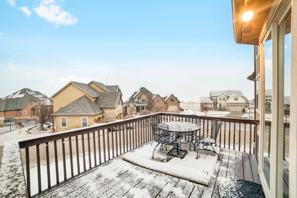 Snowy deck overlooking homes in the village blanketed with snow in winter. Table, chairs, glass door, and blue sky can also be seen in this frosted scenery.