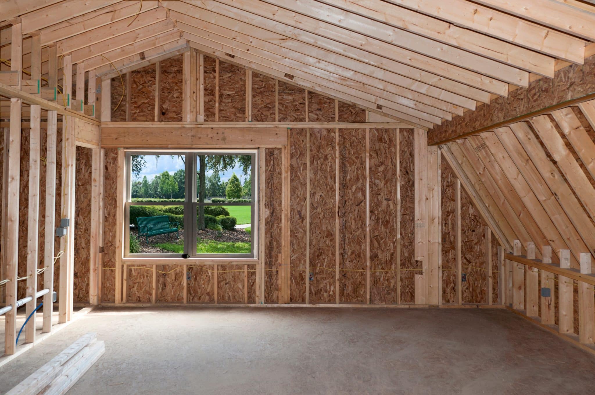 Interior view of a room under construction with wooden trusses and particle board walls, featuring a large window offering a view of a landscaped garden.