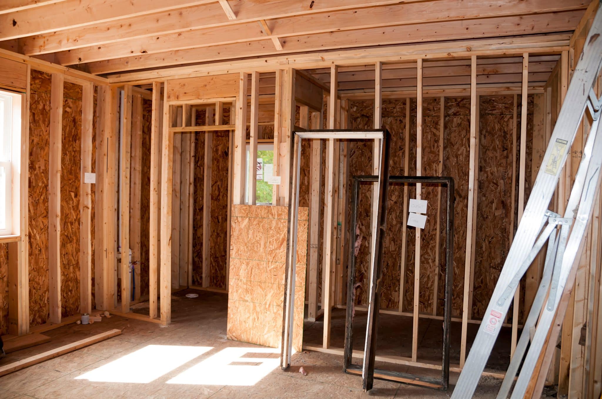 Early construction phase of a room addition showing exposed wooden framing, particle board, and the installation of plumbing within a brightly lit structure.