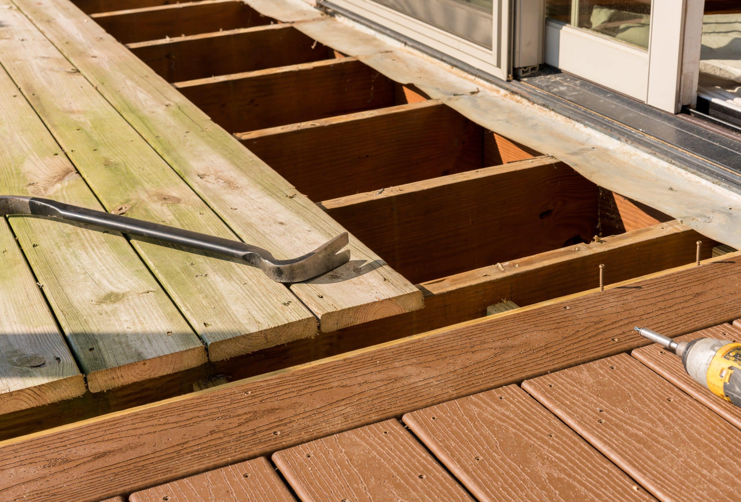 Photo of a residential decking renovation in progress, showcasing an array of decking materials. Partially disassembled wooden deck boards are visible, with new, fresh lumber laid out next to existing brown-stained planks. A crowbar and a cordless drill are on the deck, indicating ongoing work.