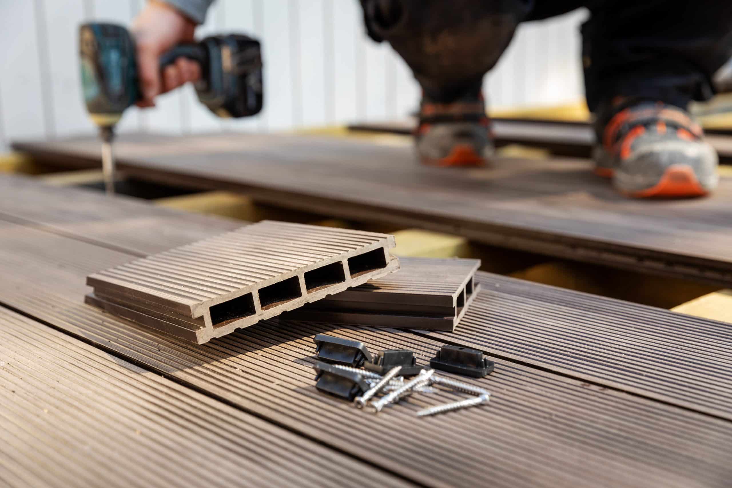 Close-up image of composite decking materials on a newly constructed deck with a person in the background using a cordless drill. In the foreground, cut cross-sections of composite deck boards reveal the internal structure, alongside a selection of metallic screws and brackets arranged on the brown textured surface.